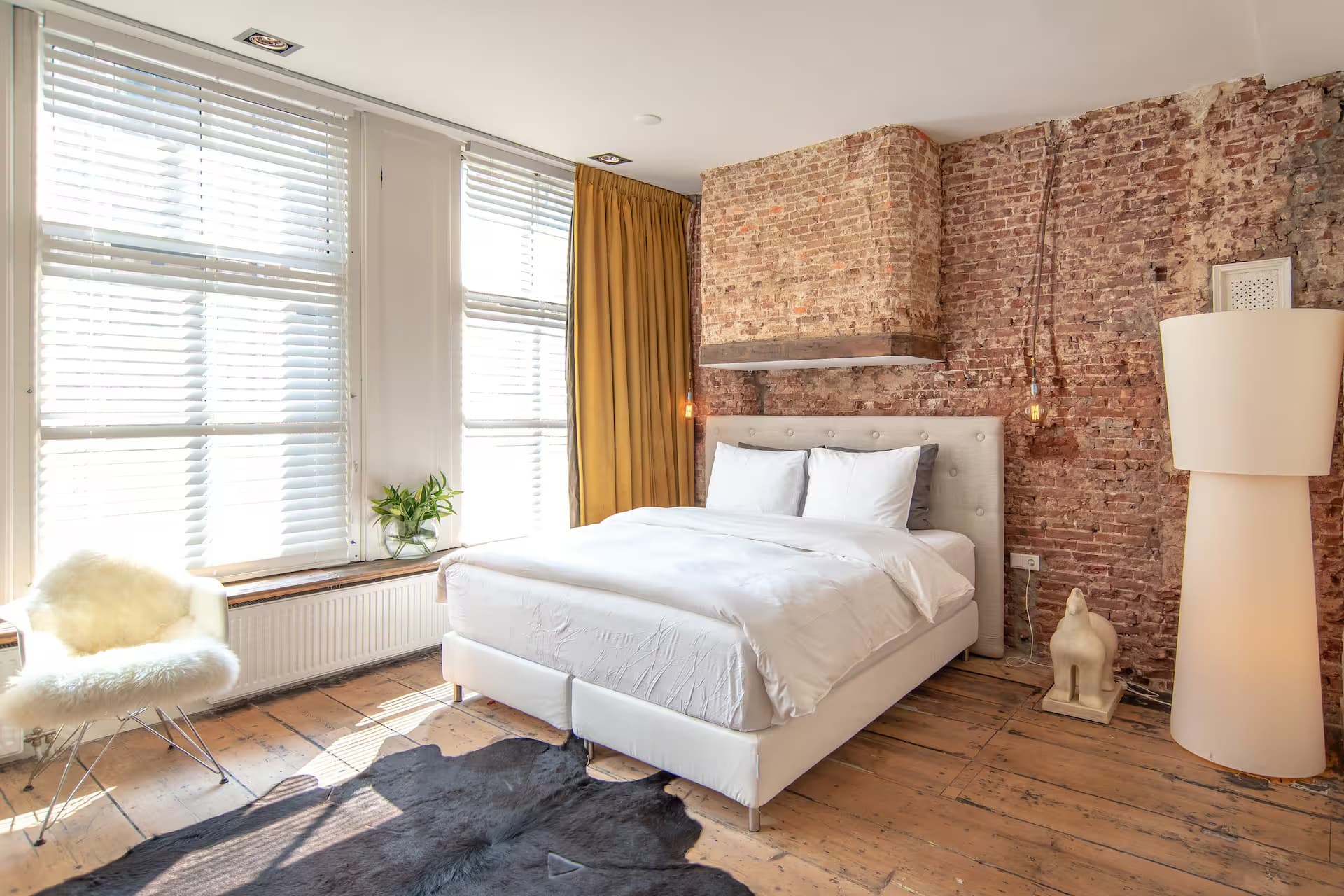 Double Fun Wolvenstraat 16 — kingsize bed tegen brick-wall met gouden velvet gordijnen, witte zonwering en cowhide-tapijt